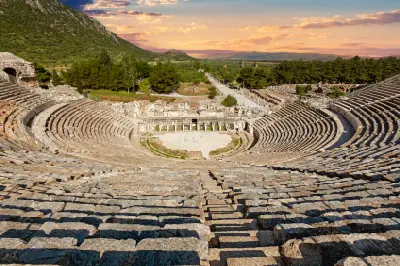 Panoramic view from the upper stone seating of the Great Theatre of Ephesus, looking across the semicircular stage area toward the ancient city and distant mountains.