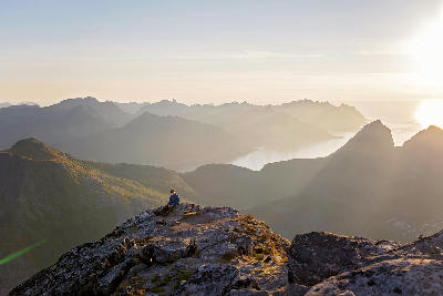 Hiker sitting on rocky summit at Grytetippen Senja Norway overlooking fjords and mountain ridges in golden sunset light.