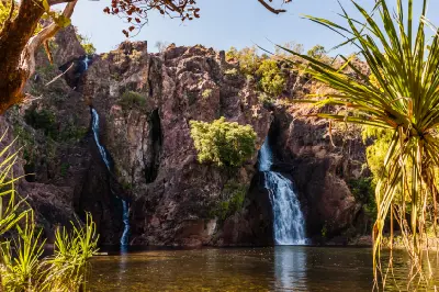 Two waterfalls descending dark rocky cliffs into a calm plunge pool, framed by pandanus leaves and tropical vegetation at Wangi Falls, Litchfield National Park, Northern Territory, Australia.