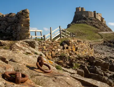 Lindisfarne Castle on a steep rocky outcrop above Holy Island's shoreline, with rusted iron mooring rings, stone walls and wooden fences in the foreground.