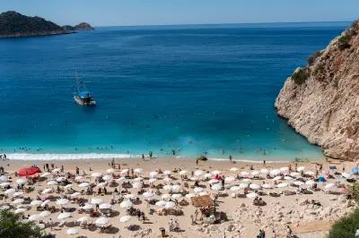 View over Kaputaş Beach, with rows of sun umbrellas, swimmers in turquoise Mediterranean water and steep rocky cliffs enclosing the cove.