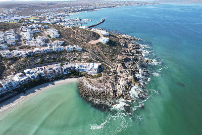 Drone view of Club Mykonos resort and marina in Langebaan, showing whitewashed waterfront buildings, rocky coastline and the turquoise waters of Langebaan Lagoon.