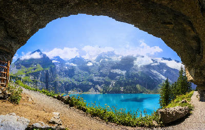 Oeschinensee seen through a rock overhang along a mountain trail, showing turquoise water and steep alpine cliffs above Kandersteg, Switzerland.