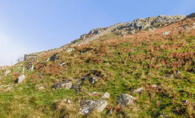 Hadrian's Wall ascending a rocky hillside at Peel Crags in Northumberland, England, surrounded by grass, bracken and scattered stones beneath a clear blue sky.