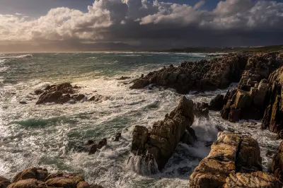 Powerful ocean waves breaking against rugged coastal rocks at De Kelders under dramatic storm clouds in the Western Cape, South Africa.