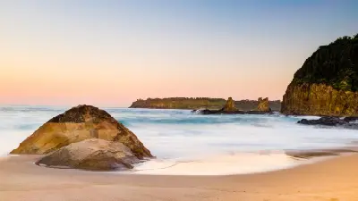 Sunset view from Jones Beach toward the Cathedral Rocks sea stacks, with smooth surf washing around large rocks on the sandy shoreline.