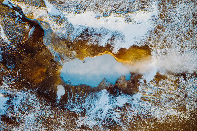 Top-down drone view of a steaming geothermal pool surrounded by yellow sulphur deposits and snow at the Seltún geothermal area in Krýsuvík, Iceland.