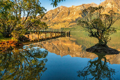 Glenorchy Wharf and willow tree reflected in calm waters of Lake Wakatipu with mountains behind, South Island, New Zealand.