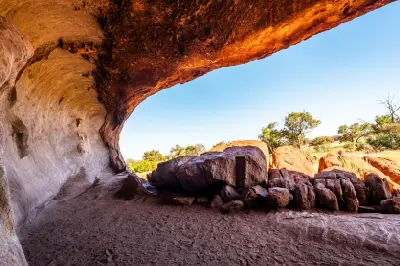View from beneath a sandstone rock shelter at Uluru, with a broad weathered overhang framing red boulders and Outback vegetation.
