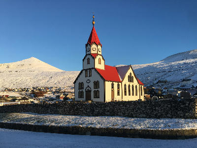 Sandavágur Church in winter, a white wooden church with a red roof and tower, surrounded by snow-covered hills and village houses in the Faroe Islands.