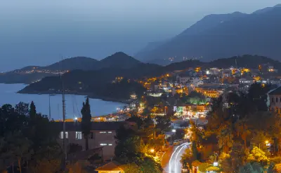 Panoramic evening view of Kaş, Turkey, showing illuminated hillside homes and winding streets beside a calm Mediterranean bay, framed by layered mountain silhouettes at dusk.