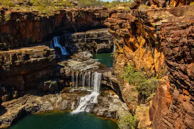 Multi-tiered Mitchell Falls cascade through a deep sandstone gorge into emerald pools surrounded by rugged orange cliffs.