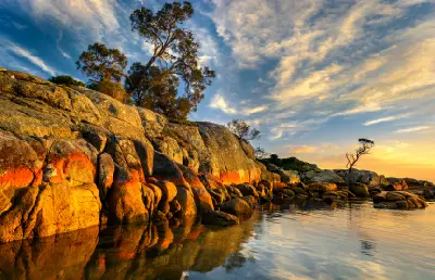 Orange lichen-covered granite rocks reflected in calm water at sunset in Tasmania’s Bay of Fires.