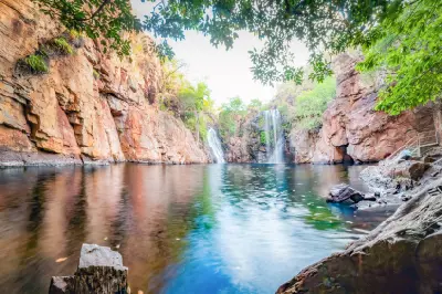 Wide view of Florence Falls with two waterfalls, a natural swimming pool, orange sandstone gorge walls and overhanging green foliage in Litchfield National Park, Australia.