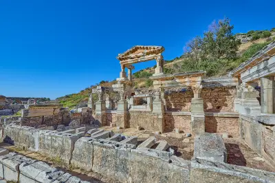 Ruins of the monumental Fountain of Trajan at Ephesus, with restored columns, carved stonework and a partially reconstructed pediment beneath a clear blue sky.