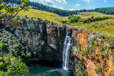 Berlin Falls dropping over orange rock cliffs into a deep plunge pool surrounded by grassland and forest in Mpumalanga.
