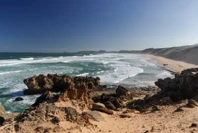 Elevated view of Brenton Beach showing rocky coastal cliffs, breaking ocean waves and a long sandy shoreline stretching along the Garden Route near Knysna, South Africa.