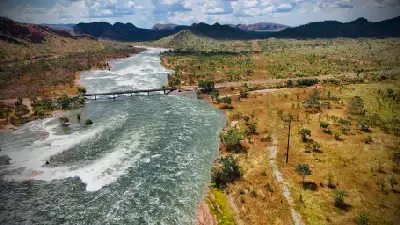 Aerial view of turbulent water flowing from Lake Argyle into the Ord River, passing beneath a bridge amid rugged Kimberley hills.