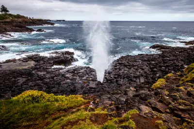 Seawater shoots vertically from the Little Kiama Blowhole as waves strike the dark volcanic rock coastline.