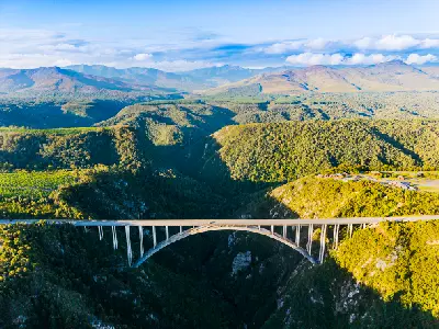 Aerial view of Bloukrans Bridge crossing a deep forested gorge in the Tsitsikamma region of South Africa, with mountain landscapes stretching across the Garden Route hinterland.