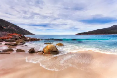 Granite boulders and sandy beach beside turquoise waves at Wineglass Bay, with forested headlands framing the ocean in Freycinet National Park, Tasmania.