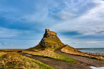 Lindisfarne Castle standing on a steep grassy and rocky hill, with winding footpaths, wooden coastal fencing and dramatic blue-grey clouds on Holy Island.