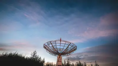 Large metal radio telescope dish above the forest canopy at Kielder Observatory, with pink clouds and blue evening sky.
