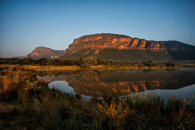 Golden sunrise light illuminating the sandstone cliffs of the Waterberg above a calm lake, with reflections of the mountains and surrounding grasslands in Entabeni Game Reserve, Limpopo, South Africa.