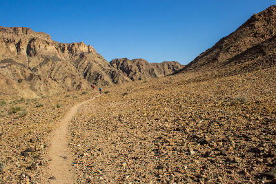 Small hikers walking along a narrow rocky desert trail beneath steep canyon mountains near Fish River Canyon in southern Namibia.