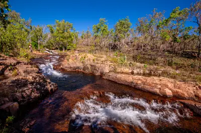 Wide view of flowing cascades and interconnected natural rock pools at Buley Rockhole, with reddish sandstone ledges, surrounding woodland and a clear blue sky.