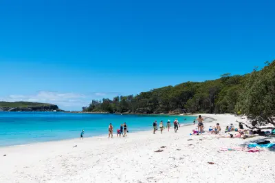 Visitors walk along the brilliant white sand of Murrays Beach beside clear turquoise water and a densely forested shoreline.