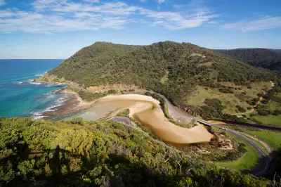 Elevated view of the Great Ocean Road curving around a sandy inlet between the ocean and forested hills near Lorne, Victoria.