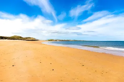 Wide sandy beach at Embleton Bay, Northumberland, with blue sea, low coastal dunes and sweeping white clouds across a bright blue sky.