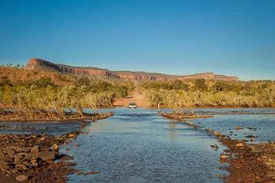 Four-wheel-drive vehicle crossing the shallow Pentecost River with the red cliffs of the Cockburn Range rising in the background.
