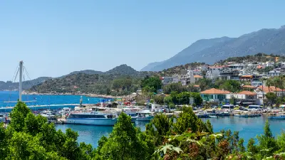 View across Kaş harbor, with yachts and excursion boats, waterfront buildings, green hills and mountains rising behind the town.