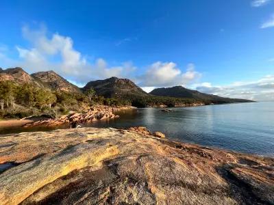 Sunlit granite rocks beside calm water at Honeymoon Bay with The Hazards rising behind under a blue evening sky, Freycinet National Park, Tasmania.