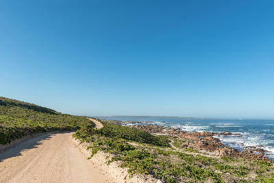 Dirt road leading through coastal fynbos to the rocky shoreline of Tietiesbaai in Cape Columbine Nature Reserve, with Atlantic Ocean views near Paternoster, South Africa.