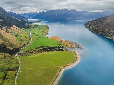 Aerial view of Lake Hāwea shoreline with farmland, winding road, and surrounding mountains, South Island, New Zealand.
