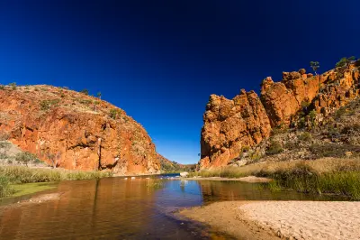 A calm waterhole of the Finke River flows smoothly between the towering, sunlit orange rock cliffs of Glen Helen Gorge under a deep blue sky.