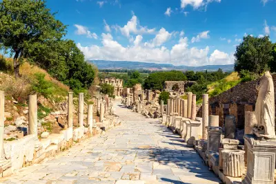 View down the column-lined Curetes Street in ancient Ephesus toward the Library of Celsus, surrounded by Roman ruins, trees and distant hills.
