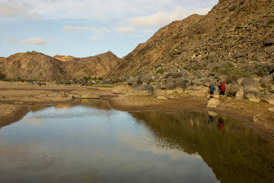 Two hikers standing beside reflective water within the rocky desert landscape of Fish River Canyon in southern Namibia.