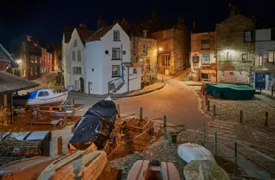 Night view of Robin Hood's Bay in North Yorkshire, showing warmly lit stone cottages, a winding village street and small fishing boats in the foreground.