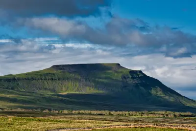 Ingleborough mountain with its distinctive flat summit and green limestone slopes beneath a dramatic blue and cloudy sky in the Yorkshire Dales.