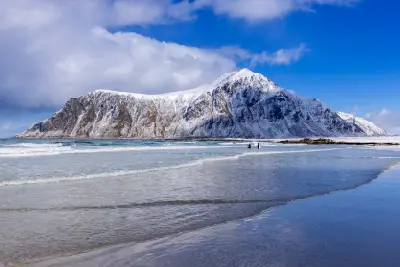 Snow-covered mountains rising above Unstad Beach on Vestvågøya in the Lofoten Islands, with winter waves along the sandy shoreline in Nordland, Norway.