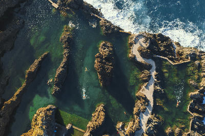 Aerial view of the Porto Moniz natural lava pools in Madeira, showing swimmers in clear Atlantic seawater separated by dark volcanic rock formations.
