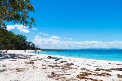 Visitors relax on the bright white sands of Murrays Beach beside calm turquoise water and a forested shoreline in Booderee National Park.