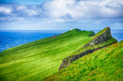 White lighthouse with red roof on a green ridge at Mykines Hólmur overlooking the Atlantic Ocean, Faroe Islands.