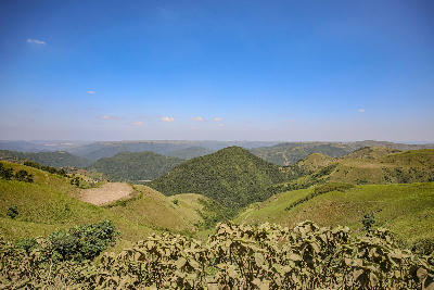 Panoramic view across the rolling hills, grasslands and rural valleys of the Valley of a Thousand Hills, with scattered farms and layered ridgelines stretching into the distance beneath a clear blue sky.