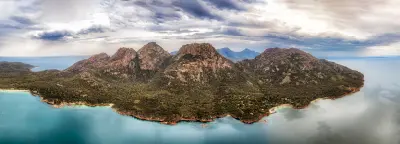 Aerial panorama of The Hazards mountain range surrounded by forest and turquoise coastal waters in Freycinet National Park, Tasmania.