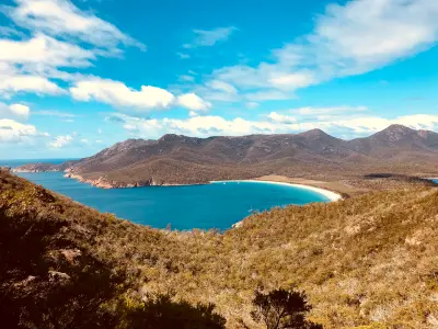 Panoramic view of Wineglass Bay with deep blue water, sweeping white-sand beach and rugged mountains in Freycinet National Park, Tasmania.